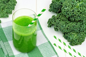 Green kale smoothie in a glass with straws on a checkered cloth against white wood