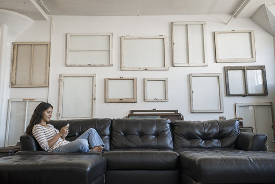 Loft decor, A wall hung with pictures in frames, reversed to show the backs, A woman sitting on a sofa using a smart phone,