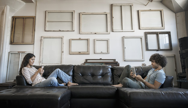 Loft decor, A wall hung with pictures in frames, reversed to show the backs, A young couple sitting on the sofa, one using a smart phone and one holding a digital tablet,