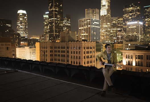 A man on a rooftop at night, playing a guitar, 