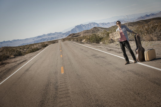 A man standing by the roadside, a hitchhiker with guitar and case, holding a sign saying Vegas or Bust, 