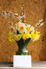 Bouquet of daffodilsin and blossoming plum branch a glass jar with a blank greeting card on a wooden table with copy space. Provence style