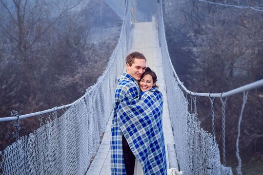 Lovely bride and groom wripped in blanket softly hugs on the wooden bridge. Honeymoon at mountains