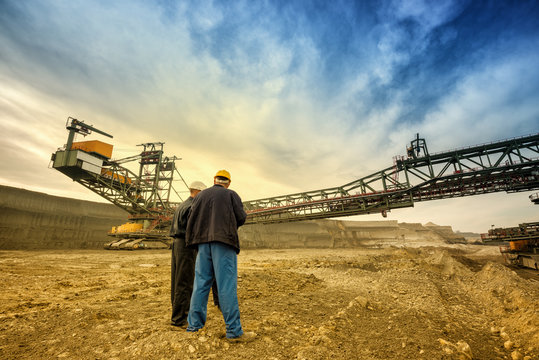 Two coal mine engineers with protective helmets standing in front of huge drill machine, chatting and watching at the digging site. Beautiful and colorful sky in the background. Rear view.
