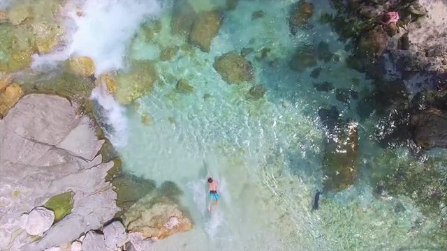 AERIAL: Young man jumping into crystal clear mountain river and starts swimming