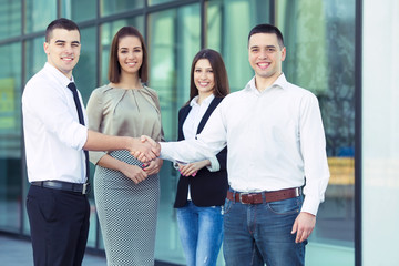 Two young businessmen are shaking hands in front of the office building. Two young businesswomen are standing next to them in the background. They are all looking at camera and smiling.