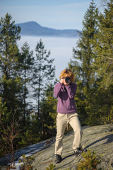 Naklejka na ściany i meble Young female photographer standing on the top of the mountain on big rock and taking picture. Front view. Trees and mountain on the background.