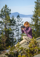 Young female photographer sitting on big rock on the mountain peak and taking picture. Trees and mountain on the background. Sunny autumn/winter day.