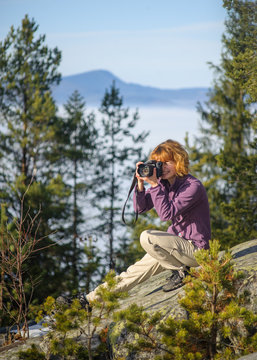 Young pretty woman photographer sitting on big rock on the mountain peak and taking picture. Trees and mountain on the background. Sunny day.