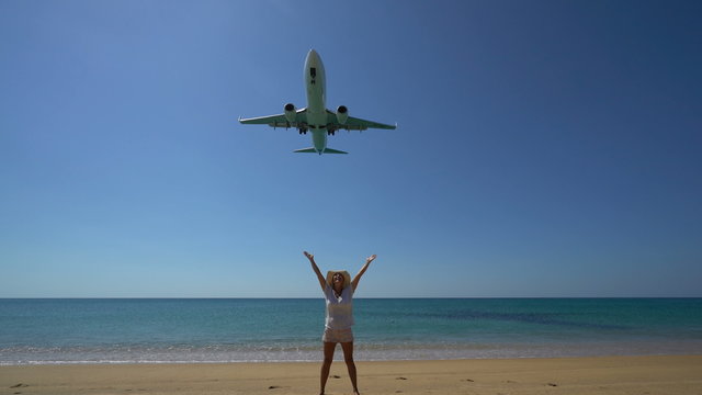 4K footage of  happy woman with raised hands on the beach with landing airplane above her. Jet engine audio included.