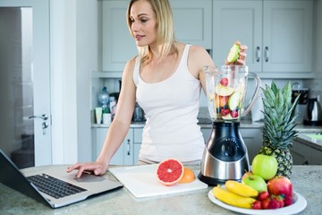 Pretty blonde woman preparing a smoothie with recipe on laptop