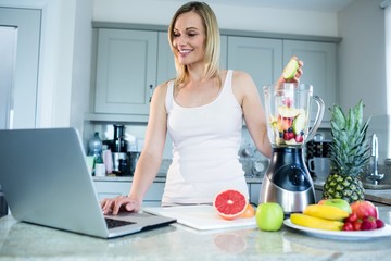 Obraz na płótnie Canvas Pretty blonde woman preparing a smoothie with recipe on laptop