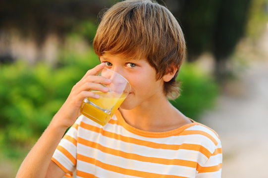 Young kid boy drinking orange juice outdoors at green background. Looking at camera. Healthy lifestyle.
