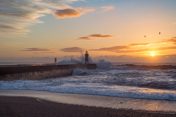 Lighthouse at sunset with rough sea, water splash around lighthouse building, flying seagulls, sunshine behind small clouds and sand beach. Porto city, Portugal.