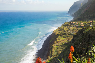 Beutiful romantic view on Madeira Island coastline - high cliffs, small village, rocks, beach, red flowers, turquoise sea water and blue sky on the horizon.