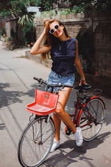 Obraz na płótnie Canvas Young pretty sensual blond girl posing outdoor with red vintage bicycle in a blue shirt in white sneakers, fashionable stylish clothes sunglasses red lipstick on her lips 