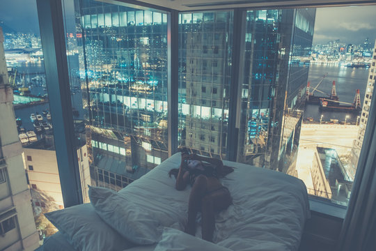 Young beautiful woman lying in the bed,making selfie or reading messages or chatting at night.Luxurious apartment with french windows,breathtaking view over harbour,skyscraper.Addiction to internet