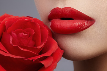 Close-up beautiful female lips with bright lipgloss makeup. Perfect clean skin, sexy red lip make-up. Beautiful visage portrait with red rose flower. Visage and cosmetics
