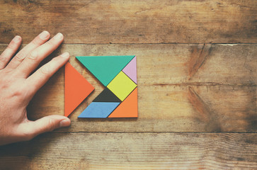 man's hand holding a missing piece in a square tangram puzzle, over wooden table.
