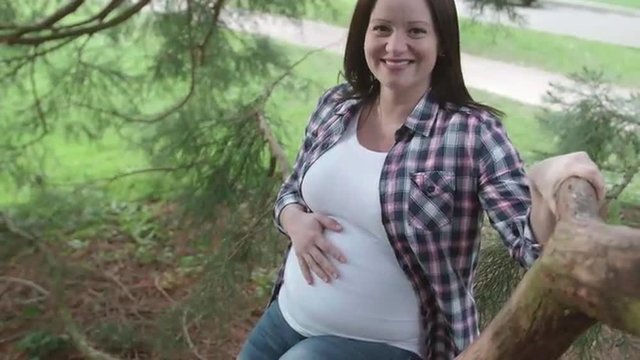 Pregnant Woman Smiling Sitting on Tree Branch Rubbing Belly