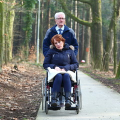 wheelchair/ old man walking with a handicapped woman in a wheelchair through the woods early spring