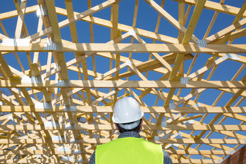 Construction worker looking at the roof of a house under construction, with a clear blue sky in the background. Horizontal format.