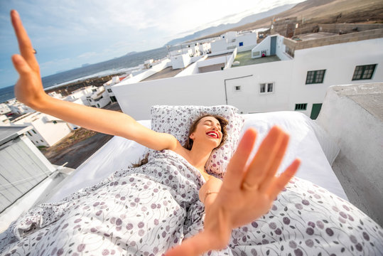 Young and cute woman straching and yawning lying on the bed on the roof top with white houses on the background. Good morning concept