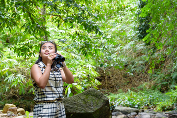 Naklejka na ściany i meble Beautiful young girl hiking is using binoculars look for birds in tropical forest near the waterfalls in Thailand