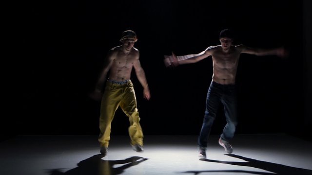Two breakdance dancers with naked torso continue dance, on black, shadow