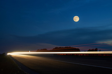 Full moon over the highway, headlights shining light lines moving cars in the landscape at dusk....