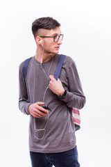Happy hipster guy in glasses with backpack using a smart phone to listen music with headphones on white background