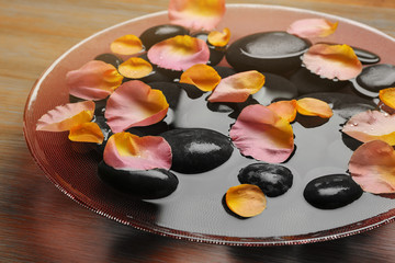 Obraz na płótnie Canvas Pink and yellow rose petals in glass bowl with water and black stones on wooden background