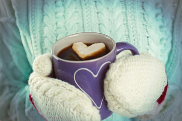 Female hands in warm mittens holding cup of hot coffee with heart marshmallow, close up