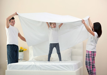Parents holding blanket above their son standing on bed in room