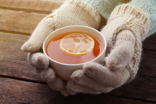 Female hands in mittens holding cup of tea on wooden background