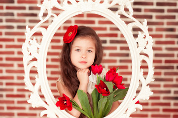 Smiling kid girl 5-6 year old holding red tulips in room over brick wall. Looking at camera. Posing in white vintage frame. Spring season. 