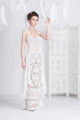 Tall and slim young bride in a luxurious lace wedding dress. She stands with her back to the viewer. She has dark hair. White feathers around her.