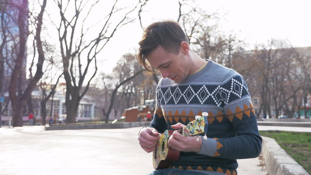 Young man playing on ukulele guitar in the park at city, 4k