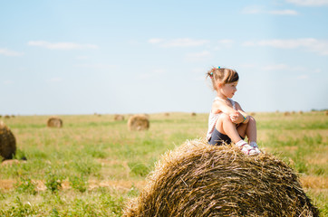 Naklejka na ściany i meble little girl sitting on a haystack in a field and watching forward