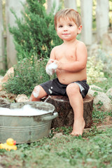 Cute baby boy 1-2 year old bathing outdoors. Child boy playing with soap foam. Childhood.