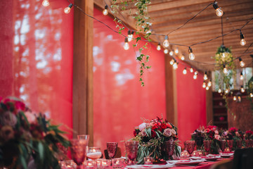 Wedding. Banquet. Marsala. In the forest on a wooden porch is served banquet table with a tablecloth color marsala and vintage chairs. On the table is floral composition and cutlery, glasses, candles