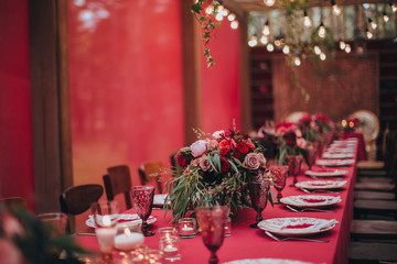 Wedding. Banquet. Marsala. In the forest on a wooden porch is served banquet table with a tablecloth color marsala and vintage chairs. On the table is floral composition and cutlery, glasses, candles