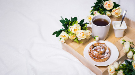 breakfast with bun, tea and yoghurt on wooden tray and flowers