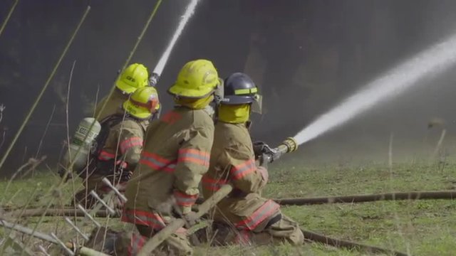 Squad of sitting firemen grasp a hose and direct water onto a house on fire, in close up