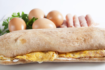 Sandwich in a bun with scrambled eggs with ham and cheese over the sour cream, set next to fresh eggs and ham.