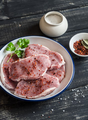 Raw pork chops and spices in an enamel bowl on a dark wooden background