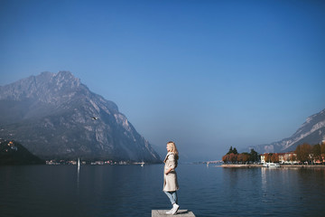 blond woman in coat standing against the background of a mountain lake Como