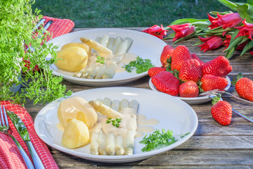 Stilleben weißer Spargel mit Kartoffeln und Sauce Hollandaise und frischen Erdbeeren, dekoriert auf altem Gartentisch mit roten Tulpen