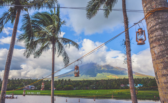 Vintage oil or kerosene lamps or lampads or lantern hanging on a rope fastened to palms against background with Mayon volcano, cloudy sky, lake. Sumilang lake, Legaspi, Philippines.