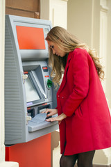 Young blonde woman in a red coat withdraw cash from an ATM in th